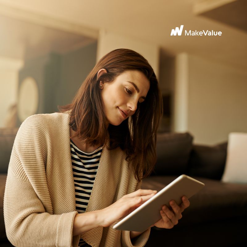 Mulher jovem sentada em casa, usando um tablet, em um ambiente confortável e iluminado, simbolizando leitura e aprendizado digital.
